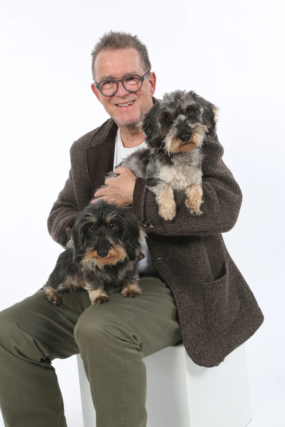 Mann mit Brille in einem braunen Blazer, der zwei kleine Hunde auf dem Schoß hält, während er auf einem weißen Hocker sitzt.