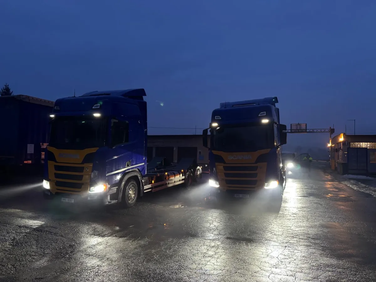 Zwei blaue Scania-Lkw mit eingeschaltetem Scheinwerfer auf einer asphaltierte Fläche bei Dämmerung.