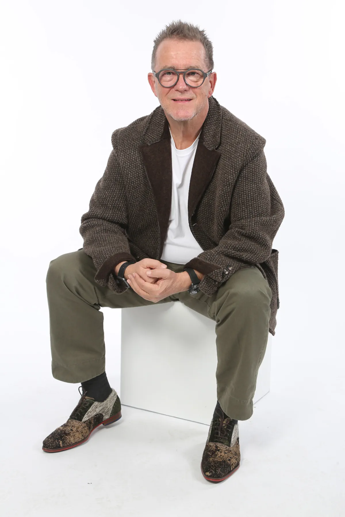 Mann mit Brille und grauen Haaren sitzt auf einem weißen Würfel, trägt einen braunen Blazer und grüne Hose mit gemusterten Schuhen.